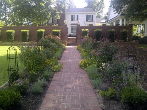 A brick pathway leads through a lush garden to a white house, framed by hedges and decorative structures.