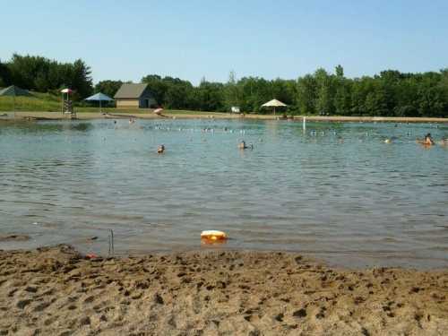 A sandy beach by a calm lake with people swimming, umbrellas, and a lifeguard station in the background.