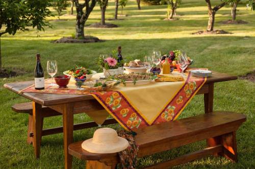 A beautifully set outdoor dining table with food, drinks, and flowers, surrounded by green trees and grass.