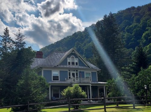 A charming blue house with a porch, surrounded by trees and mountains, under a partly cloudy sky with sunlight streaming down.