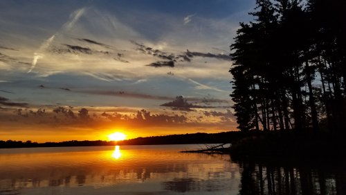 A serene sunset over a calm lake, with silhouettes of trees along the shore and colorful clouds in the sky.