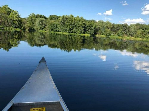 A canoe's bow on a calm lake, reflecting trees and blue sky with fluffy clouds.