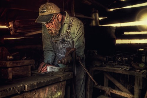 An elderly man in overalls and a cap works intently at a wooden table in a rustic workshop filled with tools.