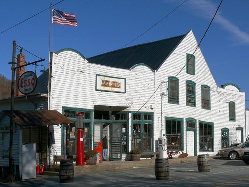 Historic white building with green trim, American flag, vintage gas pump, and barrels outside. Clear blue sky.