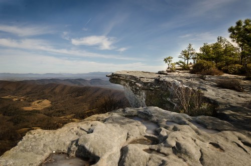 A rocky cliff edge overlooks a vast valley with rolling hills under a clear blue sky.