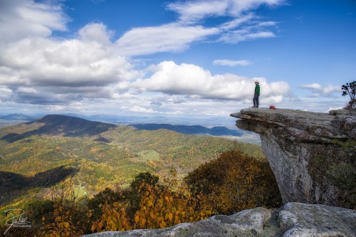 A person stands on a rocky ledge overlooking a colorful autumn landscape with mountains and clouds in the background.