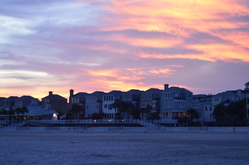 Sunset over beach houses, with vibrant pink and orange skies reflecting on a calm evening.