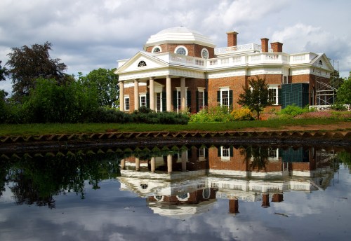 A historic brick building with a dome, reflecting in a calm pond, surrounded by greenery and cloudy skies.