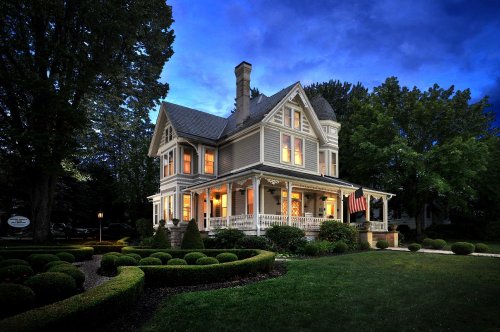 A charming Victorian-style house with a porch, surrounded by manicured gardens, illuminated at dusk.
