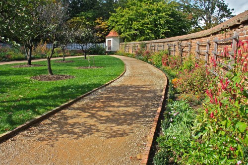 A winding gravel path through a lush garden with trees and colorful flowers alongside a brick wall.