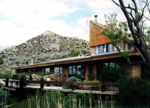 A modern house with large windows, surrounded by greenery and a mountain in the background under a cloudy sky.