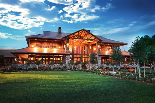 A large, modern lodge with wooden accents, surrounded by green grass and trees, under a blue sky with clouds.