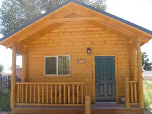 A small wooden cabin with a green door and porch, surrounded by trees and grass.