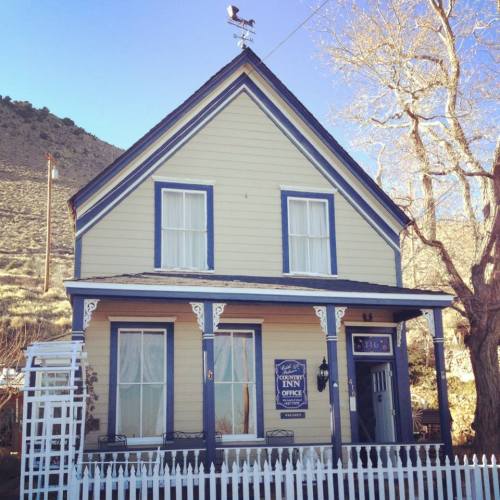 A charming yellow inn with a blue trim, white picket fence, and a weather vane on the roof, set against a mountainous backdrop.