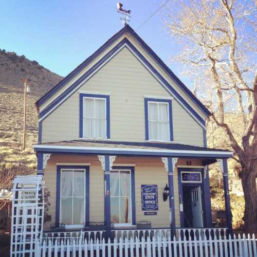 A charming yellow inn with a blue trim, white picket fence, and a weather vane on the roof, set against a mountainous backdrop.