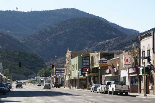 A quiet street lined with shops and cars, set against a backdrop of mountains under a clear blue sky.