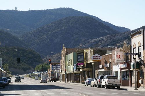 A quiet street lined with shops and cars, set against a backdrop of mountains under a clear blue sky.