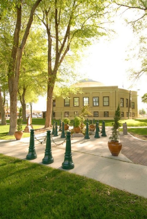 A sunny park scene featuring green trees, a building, and decorative lampposts along a pathway with potted plants.