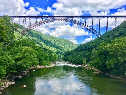 A scenic view of a river surrounded by lush green hills, with a large iron bridge arching over the water under a blue sky.