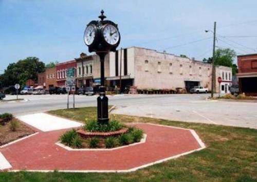 A town square featuring a large clock, surrounded by brick pathways and buildings in the background on a sunny day.