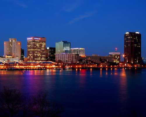 City skyline at dusk, reflecting lights on the water, with tall buildings and a clear blue sky.