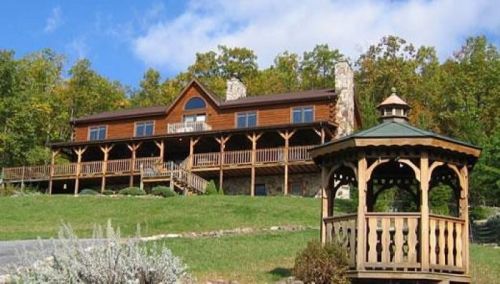 A large wooden lodge with a wraparound porch, surrounded by trees and a gazebo in the foreground.