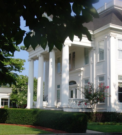 A large white house with tall columns, surrounded by greenery and flowers, under a clear blue sky.