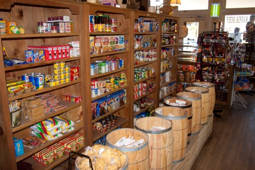 A cozy store interior with wooden shelves filled with various snacks, canned goods, and barrels for bulk items.
