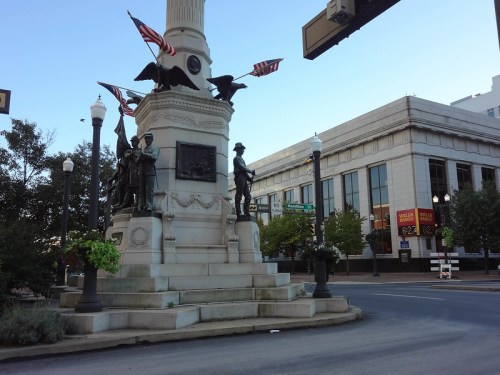 Monument with statues and flags, located at a street intersection in a city, surrounded by trees and buildings.