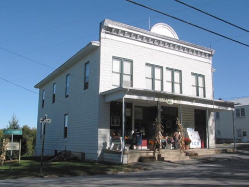 A white, two-story building with green trim, featuring a porch and people sitting outside on a sunny day.
