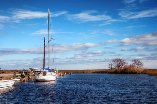 A sailboat docked by a calm waterway, with blue skies and wispy clouds above, surrounded by marshland.