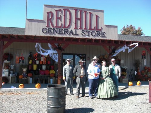 A group of people in vintage costumes stands in front of the Red Hill General Store, decorated for Halloween.