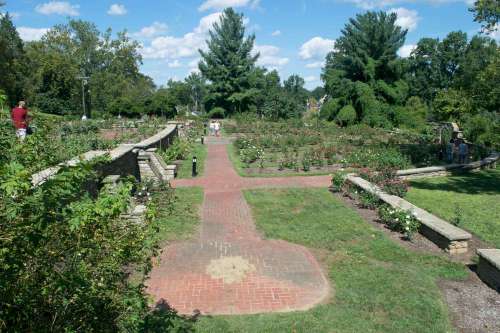 A scenic garden with brick pathways, lush greenery, and blooming flowers under a blue sky with scattered clouds.