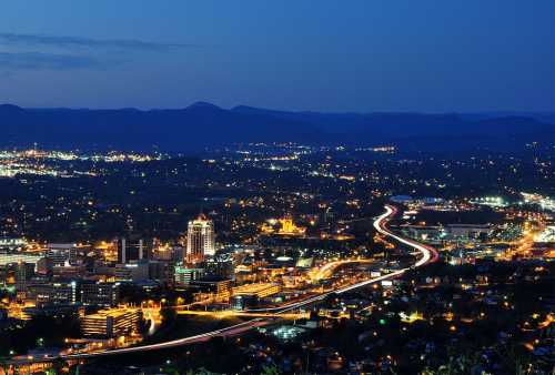 A panoramic view of a city at night, with illuminated buildings and winding roads under a twilight sky.