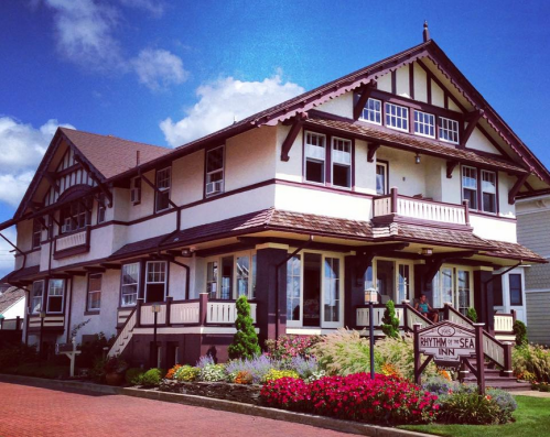 A charming, two-story inn with a decorative facade, surrounded by colorful flowers and a clear blue sky.
