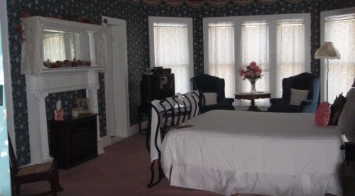 Cozy bedroom with floral wallpaper, a large bed, two armchairs, a fireplace, and bright windows.