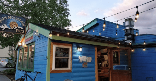 A colorful blue building with string lights, housing a pie shop, surrounded by trees and a bicycle parked outside.