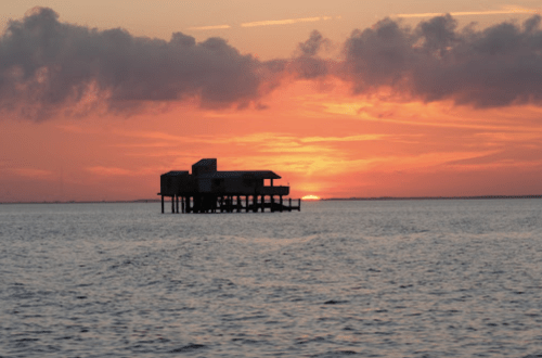 A silhouetted structure on stilts in the water against a vibrant sunset sky with clouds.