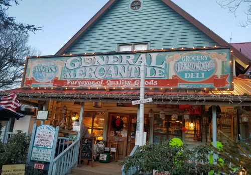 A charming general mercantile store with a colorful sign, featuring groceries, hardware, and a deli. Festive lights adorn the exterior.