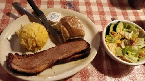 A plate with sliced brisket, cheesy rice, a roll, and a side salad with cucumbers and croutons on a checkered tablecloth.