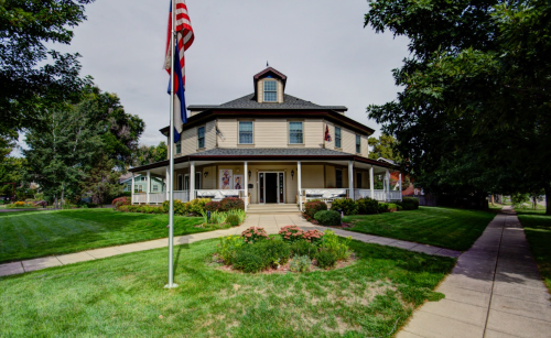 A large, round, two-story house with a porch, surrounded by green grass and trees, with flags in front.