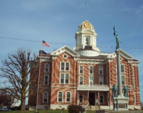 Historic brick courthouse with a clock tower, flanked by an American flag and a statue on a pedestal. Clear blue sky.