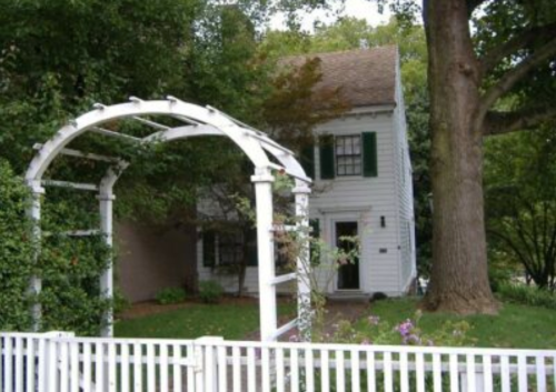 A white house with green shutters, surrounded by trees and a white picket fence, featuring an archway covered in greenery.