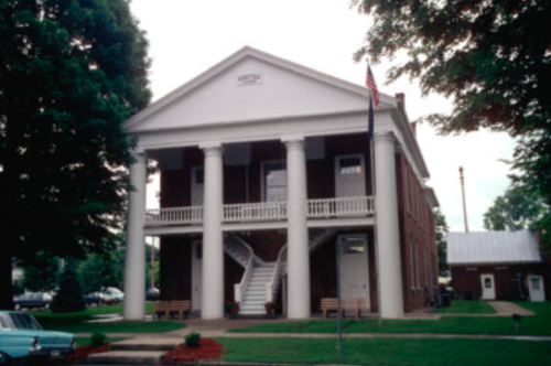 Historic building with white columns, a balcony, and an American flag, surrounded by greenery and benches.