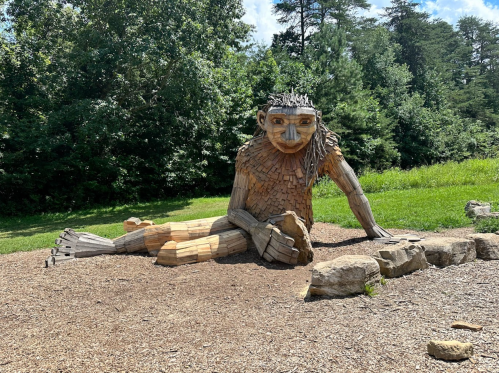 A large wooden sculpture of a troll-like figure sitting on the ground in a grassy area surrounded by trees.