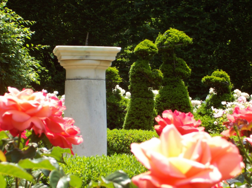 A stone pedestal surrounded by vibrant roses and neatly trimmed topiary figures in a lush garden.
