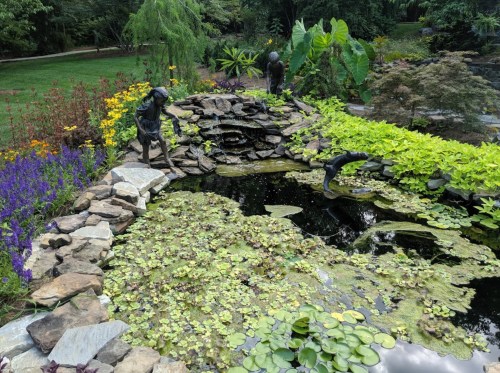 A serene garden scene featuring a pond with lily pads, surrounded by colorful flowers and stone sculptures.