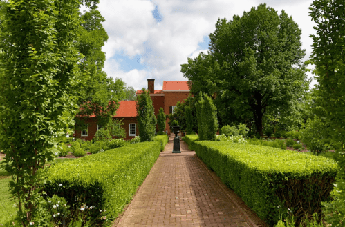 A brick pathway lined with hedges leads to a red-roofed house surrounded by lush greenery and trees.