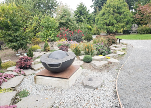 A round stone sculpture sits on a pedestal in a landscaped garden with colorful flowers and greenery.