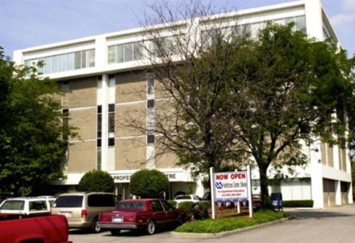 A multi-story office building with a "Now Open" sign and parked cars in front, surrounded by trees.